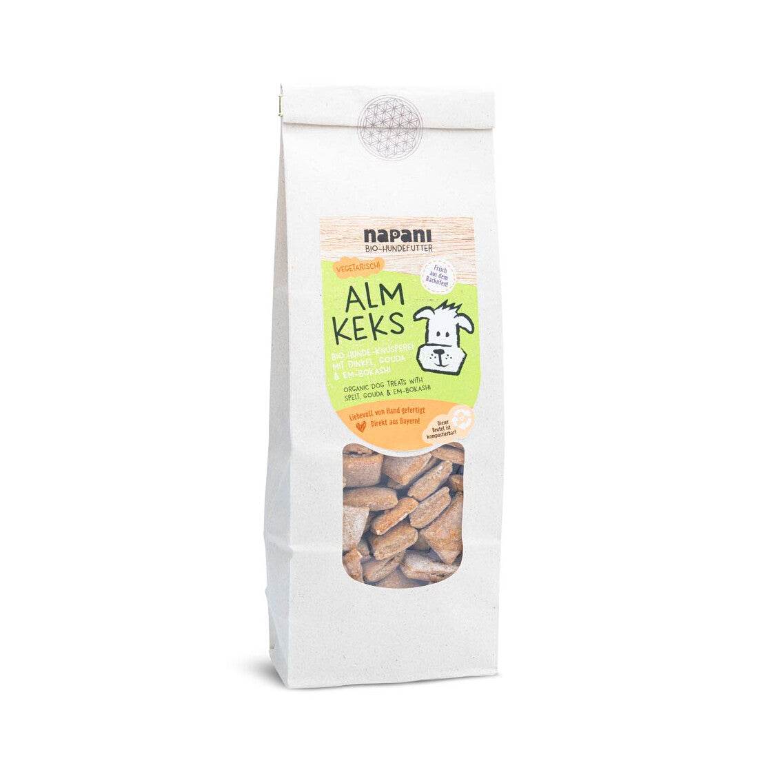 Eine Packung Bio "Alm-Kekse" für Hunde auf einem rustikalen Holztisch, umgeben von natürlichen Zutaten wie Bio-Dinkel, Gouda-Käse und anderen natürlichen Elementen. Die Verpackung ist umweltfreundlich und aus kompostierbaren Materialien.